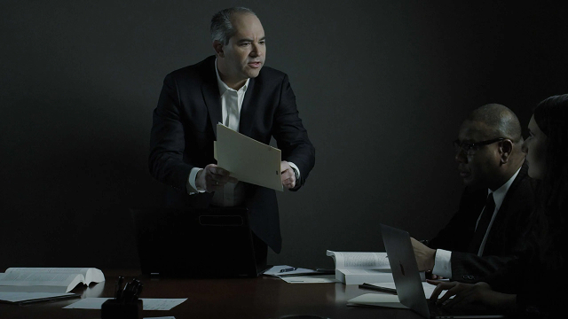MAL-in-a-Meeting-scaled 1 A man in a suit presents a folder during a dimly lit business meeting