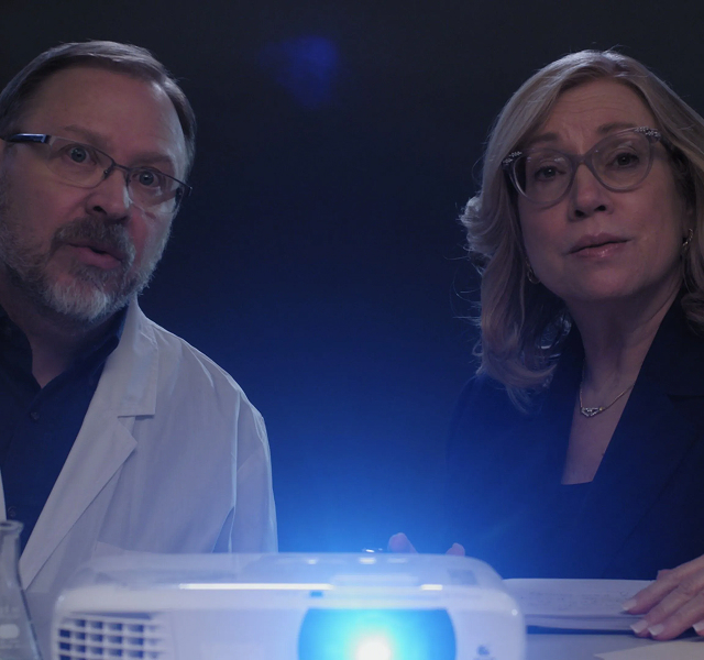 SOCwExpert 1 Two scientists in lab coats and glasses look at a glowing projector on a table